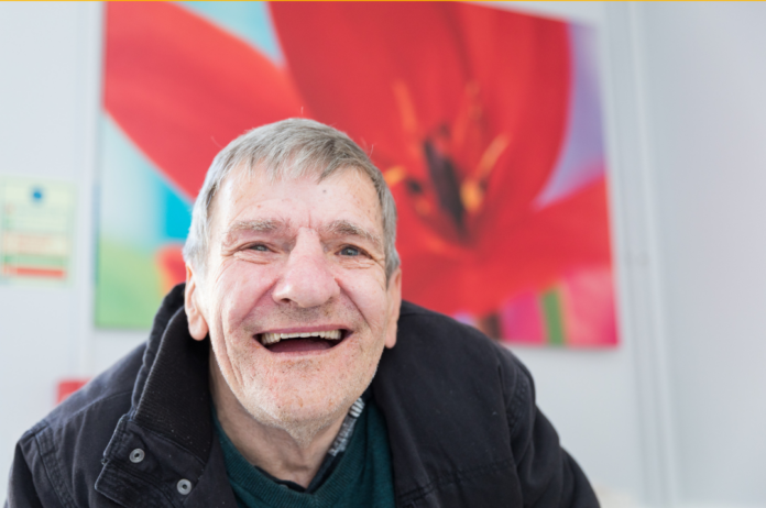 A man with grey hair smiling at the camera, in front of a bright painting of a flower.