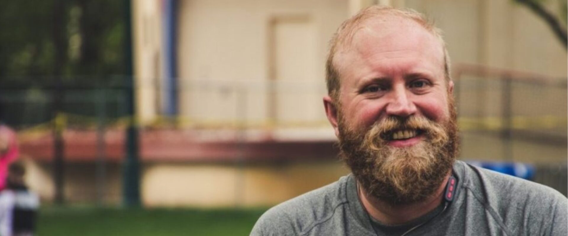Bearded man wearing a grey top, smiling outdoors.