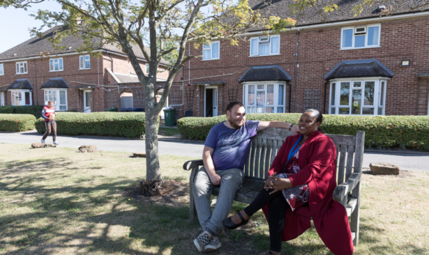 Two people sat outside red brick houses on a bench under a tree, having a conversation.