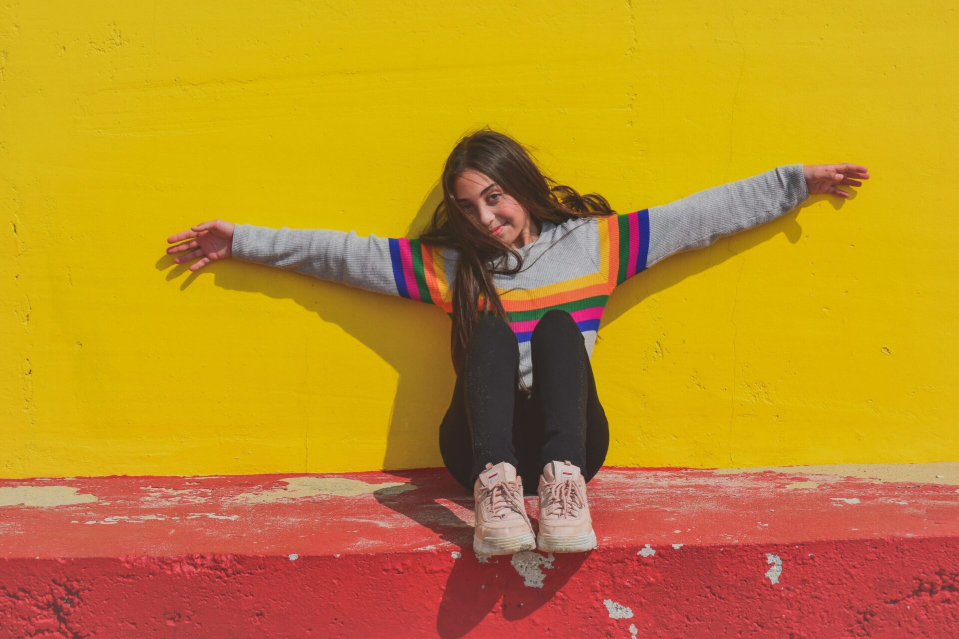 Smiling teen girl in a rainbow jumper in front of a yellow wall.