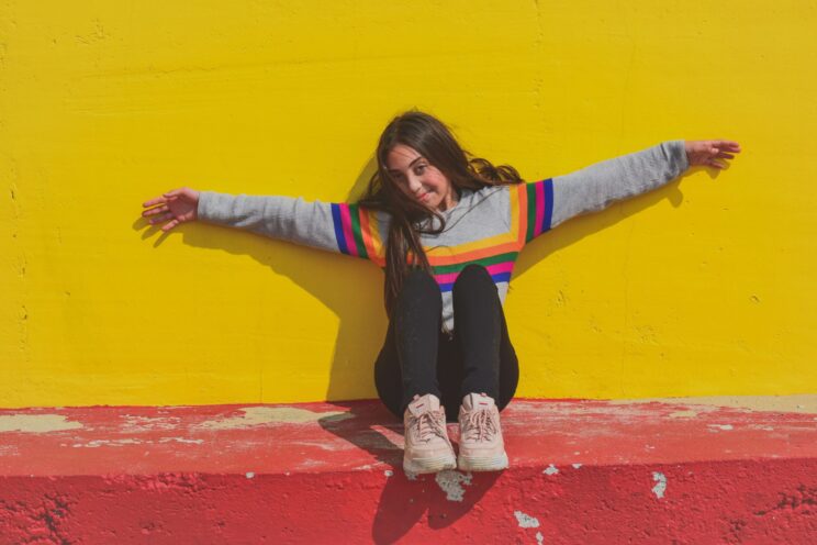 Smiling teen girl in a rainbow jumper in front of a yellow wall.