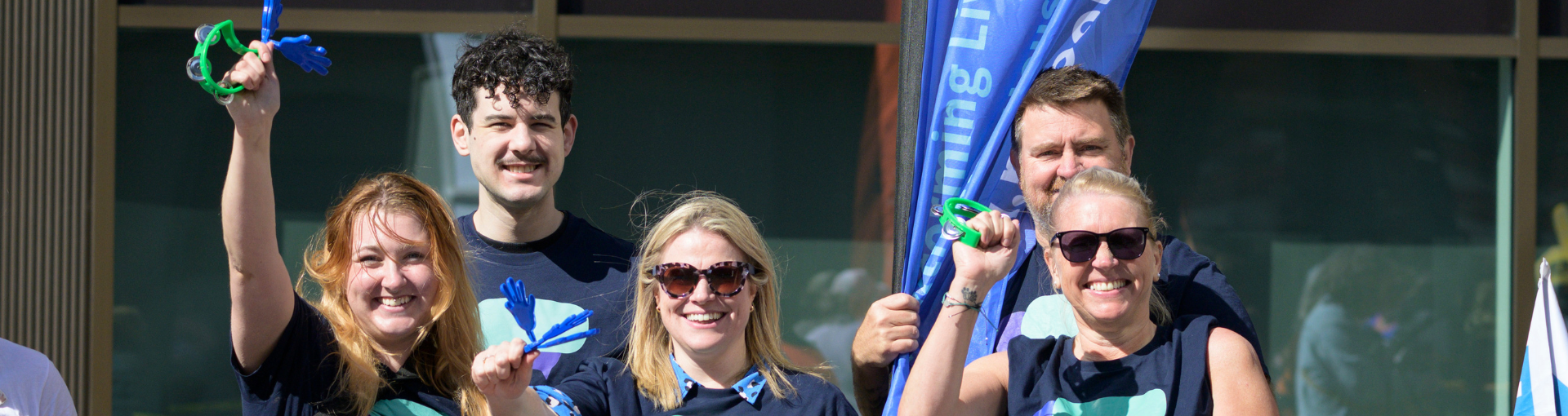 Smiling fundraisers celebrating at an outdoor charity event, holding clappers and a branded flag.