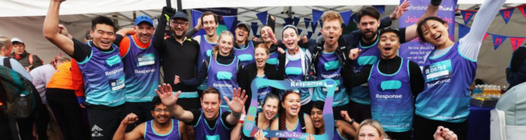 Group of marathon runners in blue “Response” charity vests celebrating together at an outdoor race event, cheering and holding a “#BeTheResponse” photo frame.