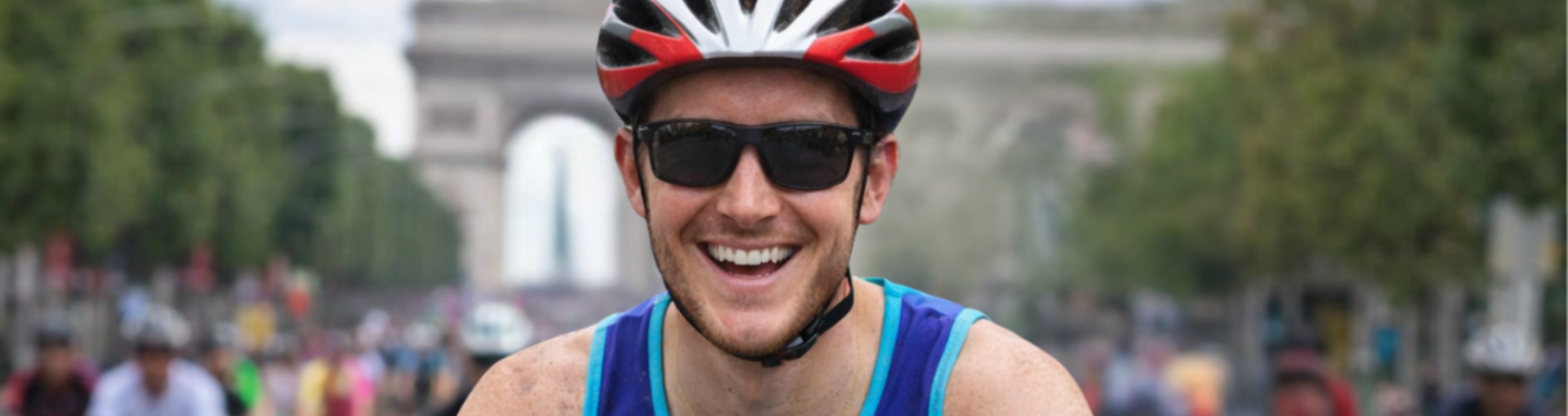 A man in a cycling helmet and sunglasses grins broadly at the camera during a charity cycle event, with a grand archway and crowds of participants visible in the background.