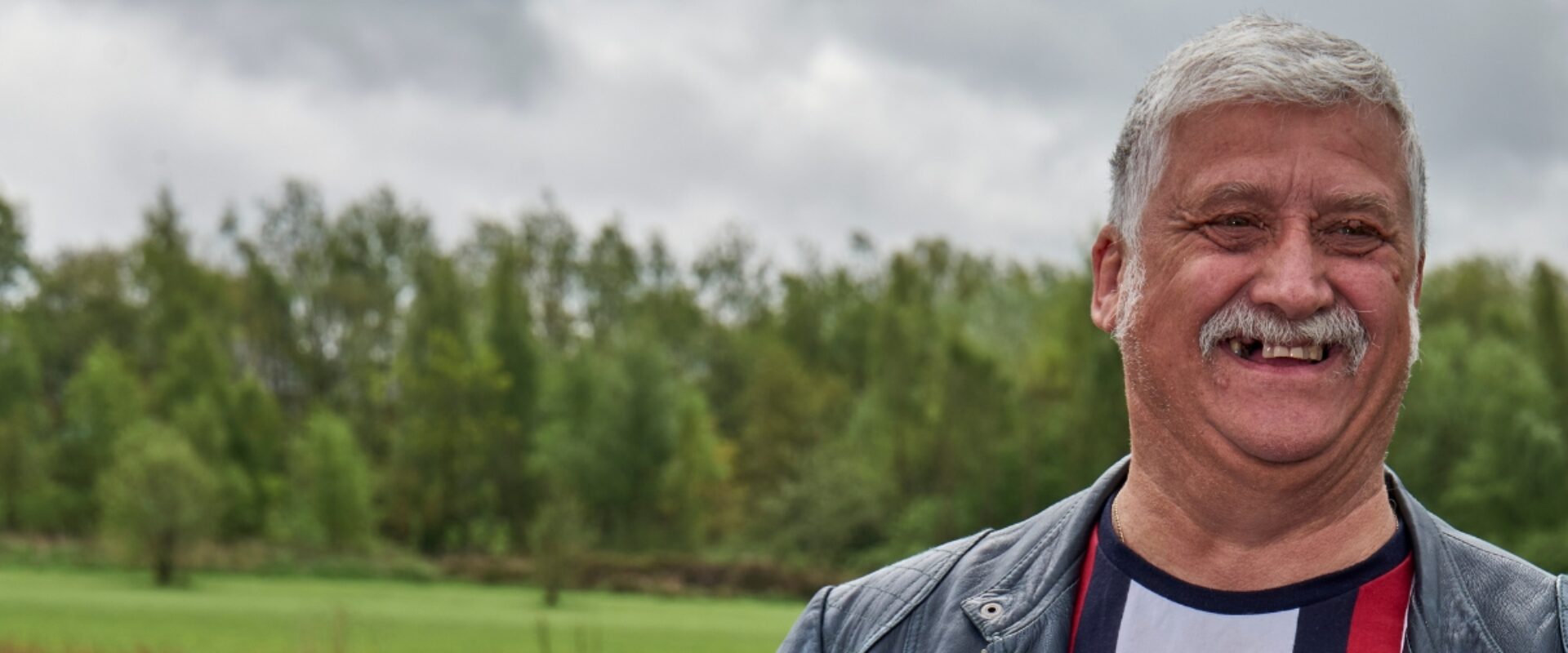 Man smiling outdoors in front of a field and green trees.