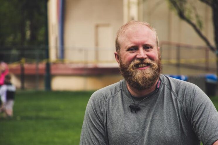Bearded man wearing a grey top, smiling outdoors.