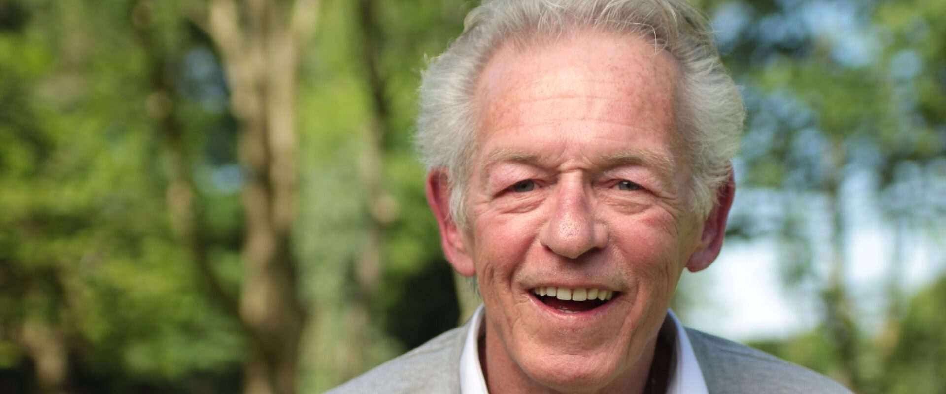 Elderly man in grey jumper and white undershirt, smiling outdoors.