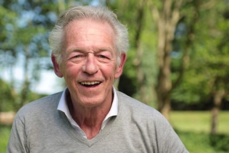 Elderly man in grey jumper and white undershirt, smiling outdoors.