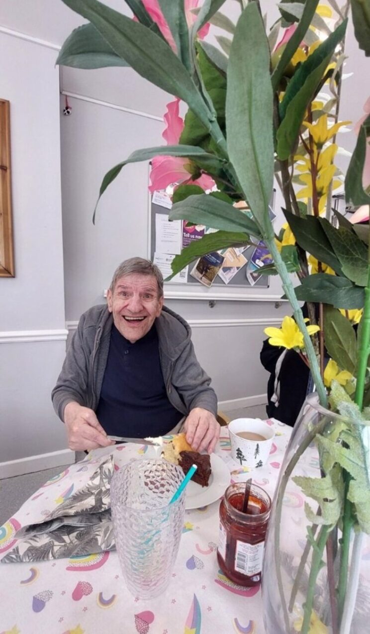 Elderly man in grey hoodie smiling indoors next to flowers.