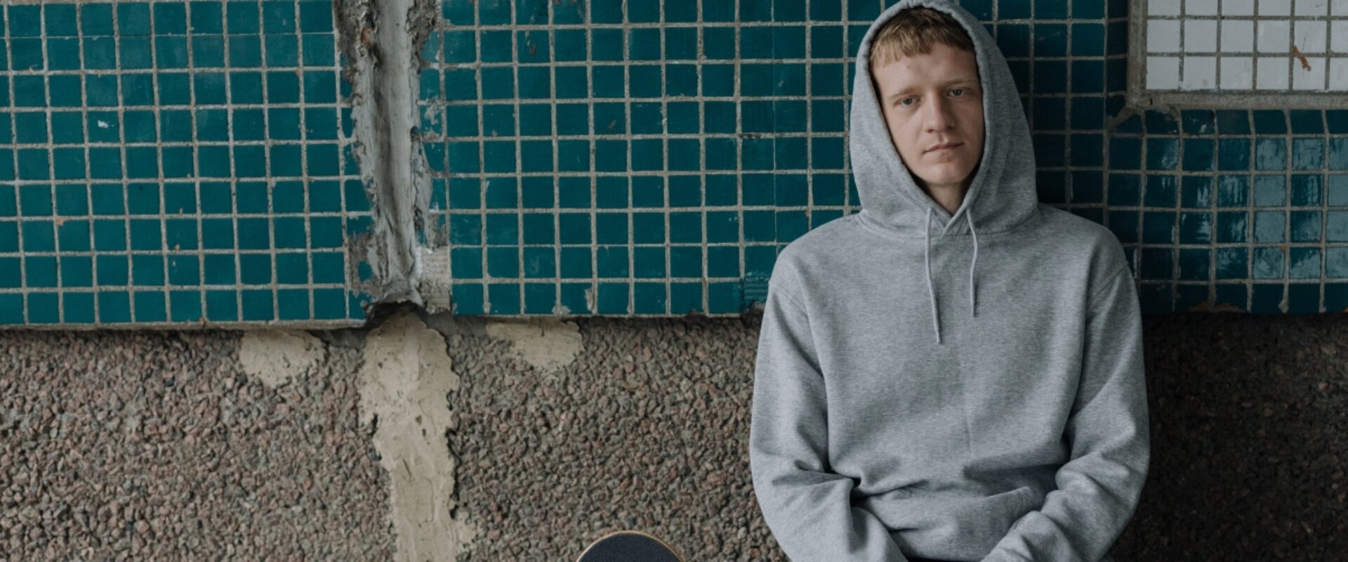 Young man sitting outside looking glum, in grey hoodie with hood pulled up.