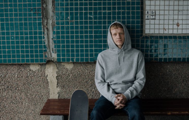 Young man sitting outside looking glum, in grey hoodie with hood pulled up.
