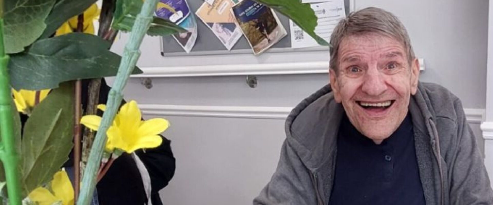 Elderly man in grey cardigan smiling indoors next to flowers.