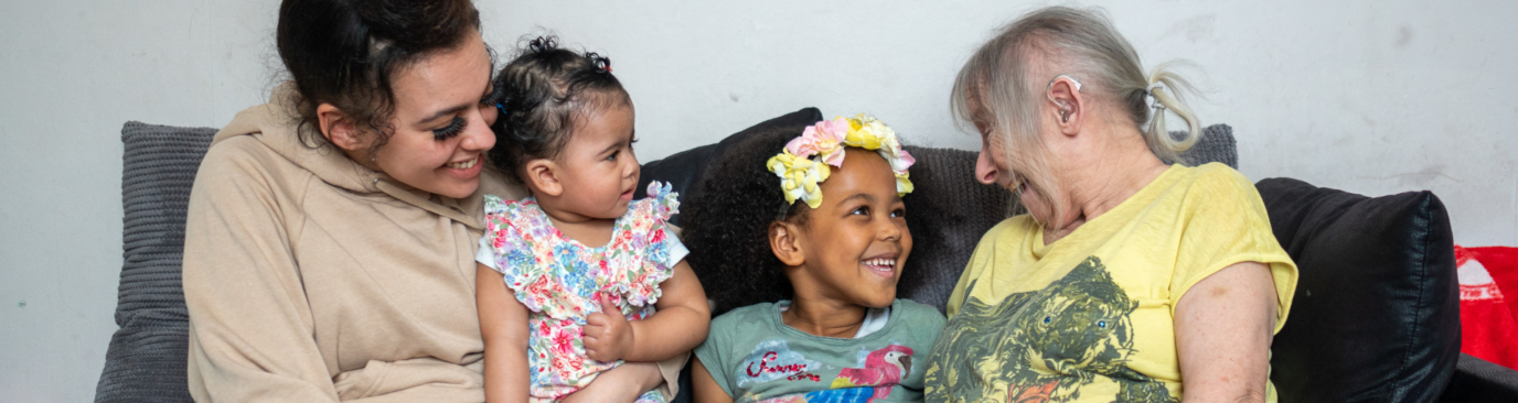 A young woman and an older woman sit together on a sofa with two young girls, all smiling and sharing a joyful moment together.