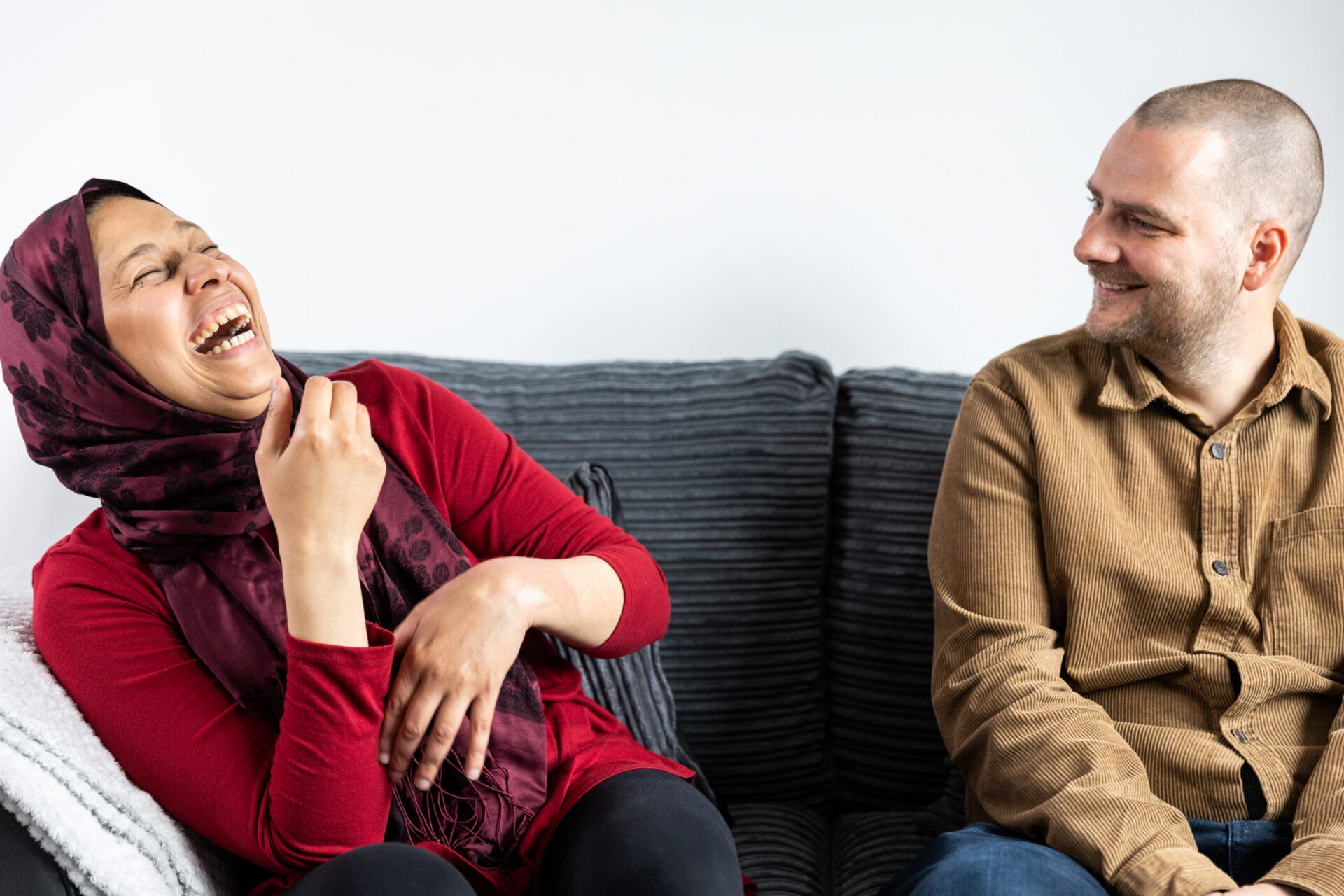 A woman wearing a red head scarf laughs and sits on a grey sofa next to a smiling man wearing a brown shirt.