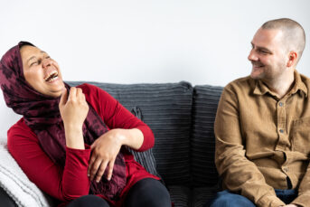 A woman wearing a red head scarf laughs and sits on a grey sofa next to a smiling man wearing a brown shirt.