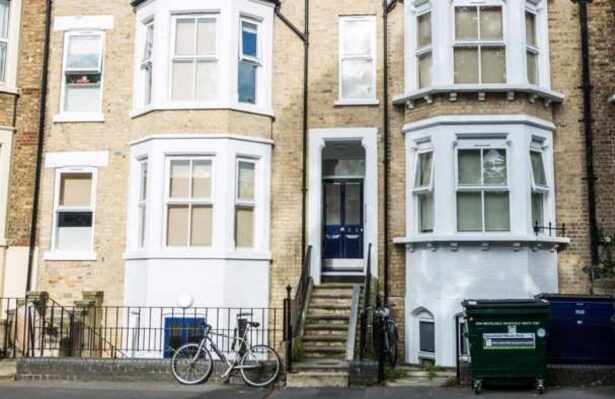 Brick house with stairs leading up to the front door. Windows are outlined with white paint and the house sits behind a black fence.