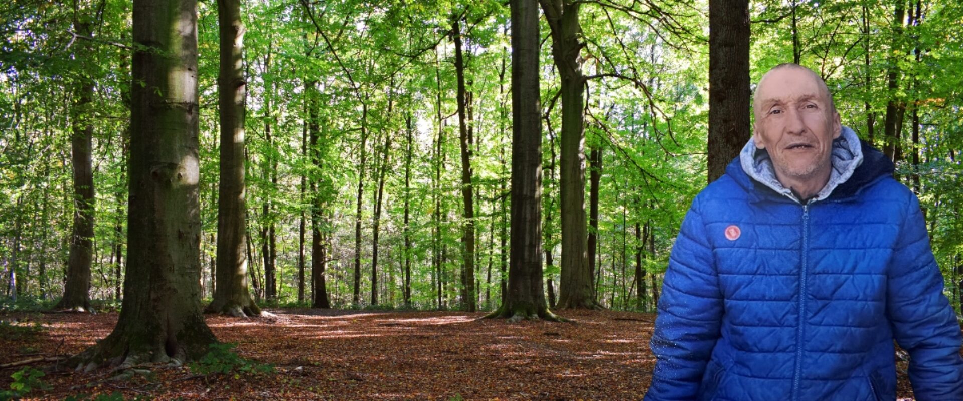 Elderly man in blue coat stood in the woods smiling.