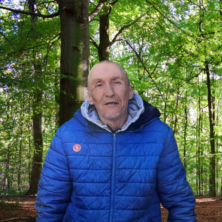 Elderly man wearing a blue coat, stood outdoors in the woods smiling.