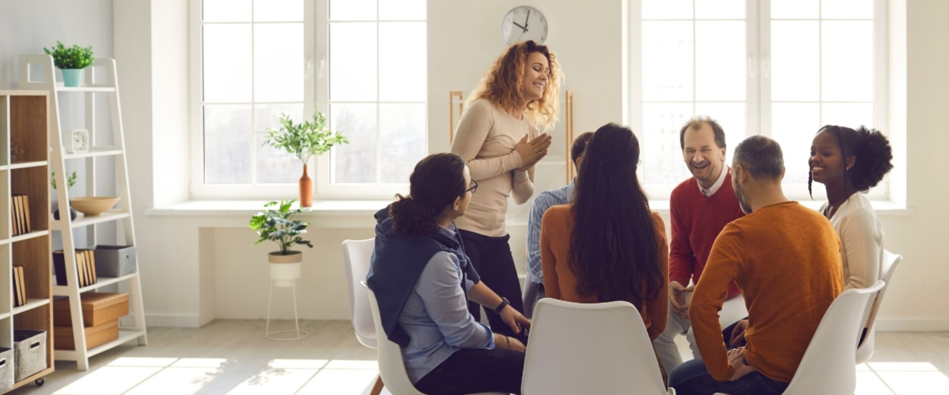 A group of people around a table studying together. There is a bookshelf to one side and a woman stands as if to share with the group.