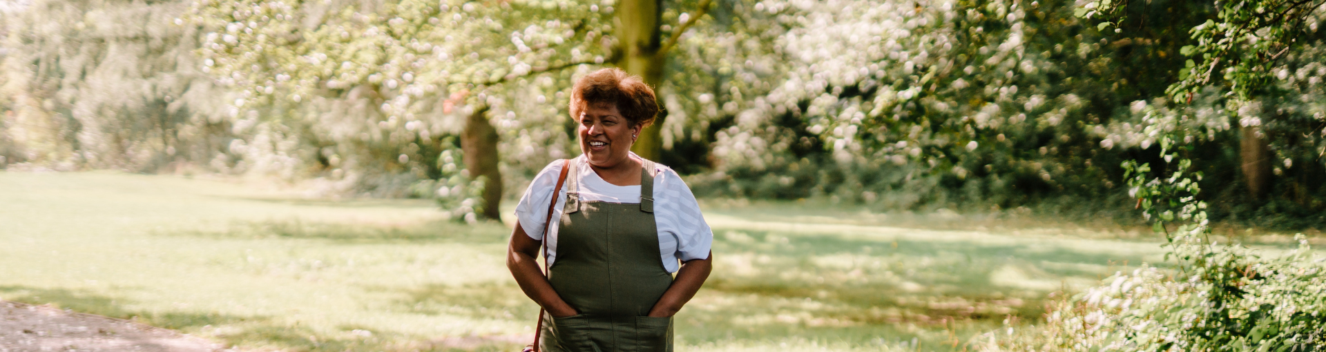 A woman in a green overalls smiles contentedly while standing in a sunlit garden surrounded by trees.