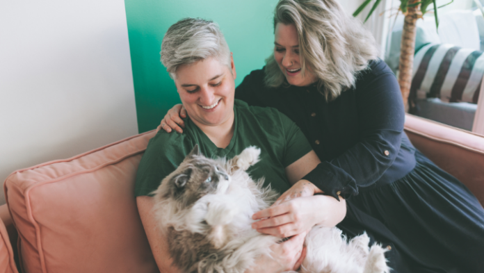 Two people sitting together on a pink sofa, laughing as they fuss over a fluffy cat.