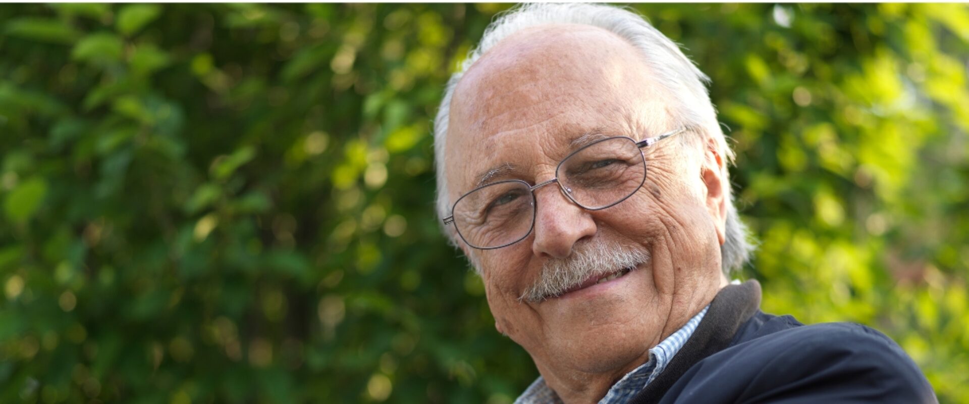 elderly man with glasses, sat outside smiling.