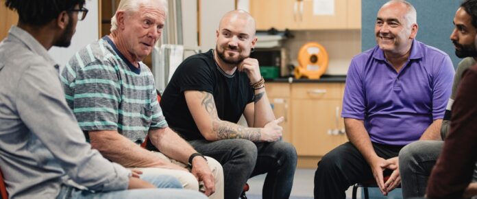 A group pf men sitting together in a kitchen having a conversation.