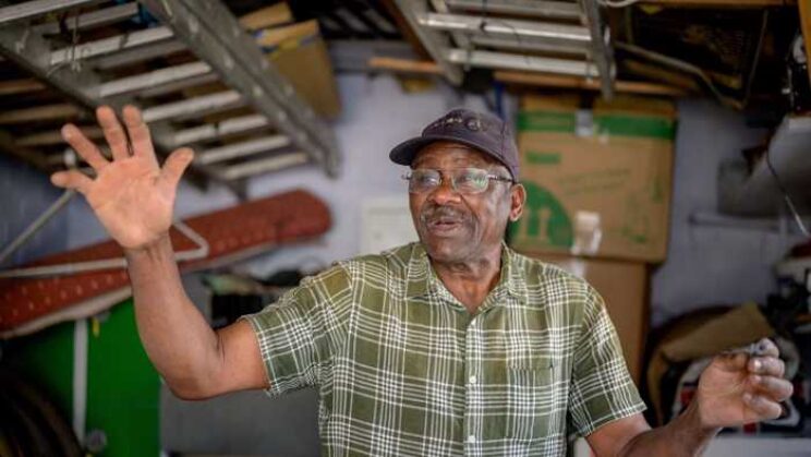 elderly man with glasses, in black cap and green striped shirt raising his hand.