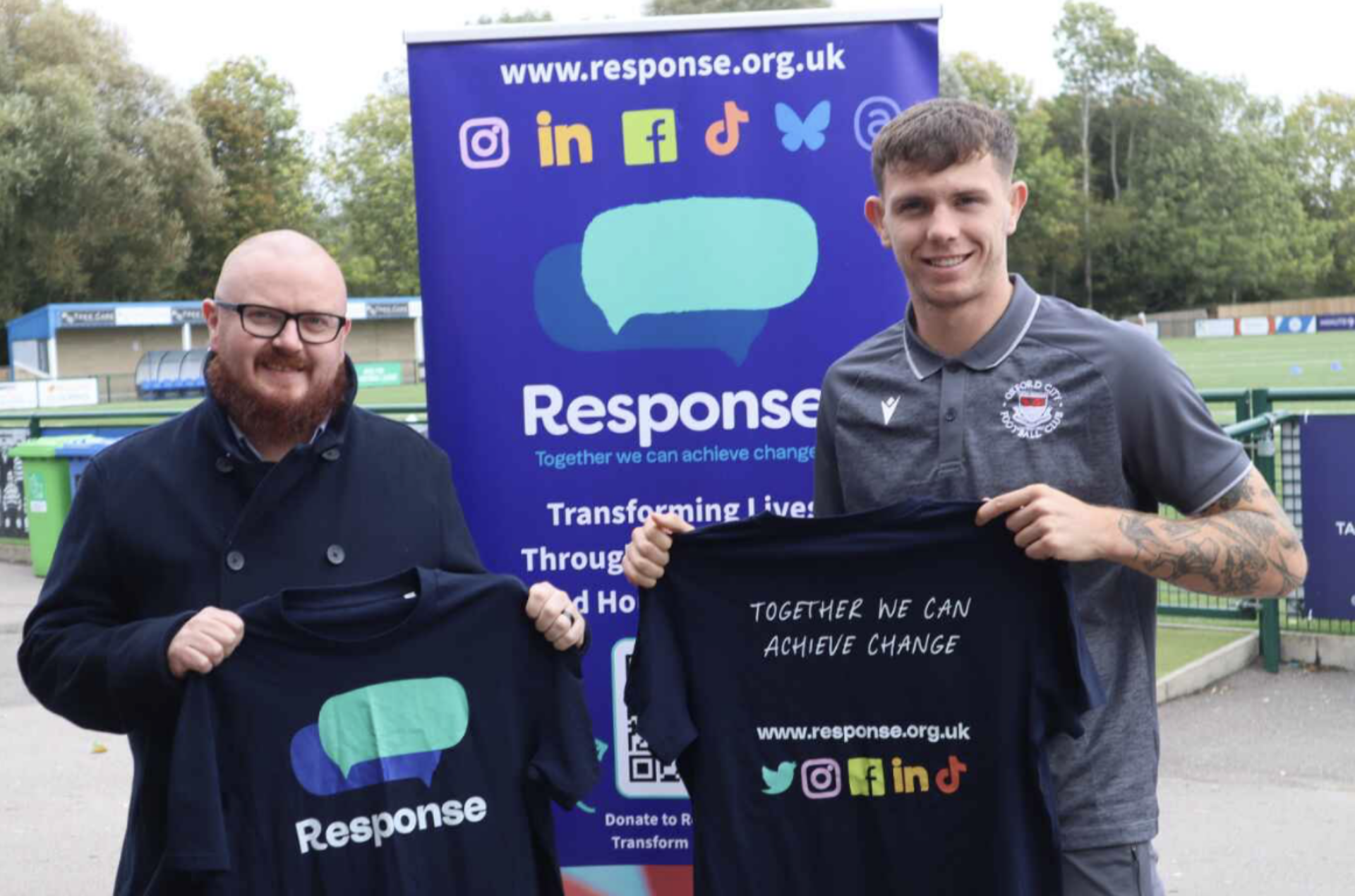TotalQ Managing Director Martin Spackman with Oxford City FC centre-half Charlie Wiggett. They stand in front of the Oxford City FC pitch holding up Response-branded t-shirts.