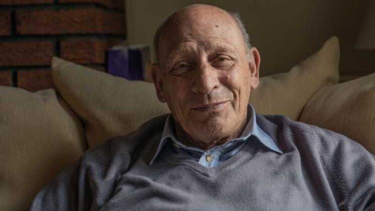 elderly man in grey jumper and blue undershirt smiling indoors.