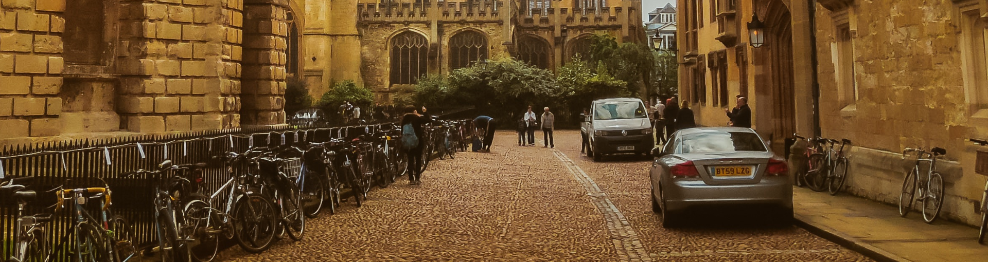 Photo of a street in Oxford by the Radcliffe Camera, an old yellow stone building, with parked cars and bicycles.