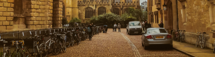 Photo of a street in Oxford by the Radcliffe Camera, an old yellow stone building, with parked cars and bicycles.