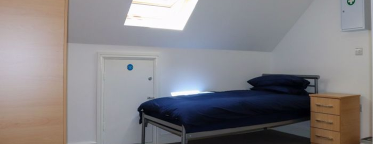 A clean, simply furnished single bedroom with a skylight, navy bedding, and a wooden bedside chest of drawers.