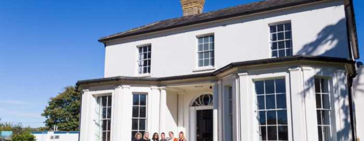 A large white Georgian-style house under a bright blue sky, with a small group of staff or residents standing together outside the entrance.
