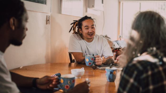 Three people sit around a kitchen table sharing drinks from colourful mugs, smiling and talking together in a warm, relaxed home setting.