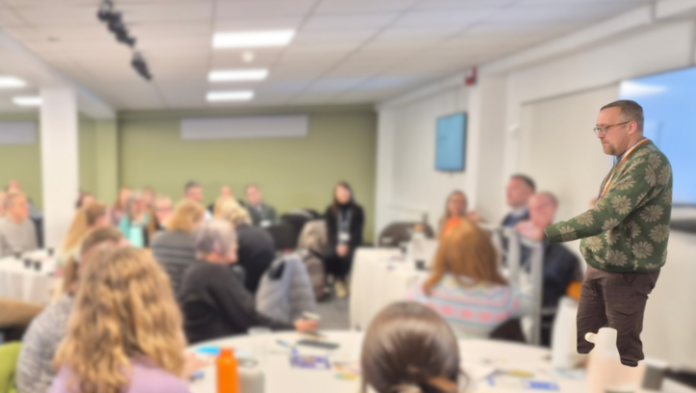 Chris Brown, Lived Experience and Involvement Coordinator at Response addresses a large group of seated attendees in a bright conference room, with a presentation screen visible in the background.