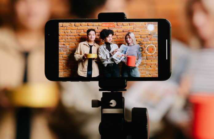 Three people are filmed on a smartphone mounted on a tripod, holding products and smiling in front of a brick wall, in a content creation setting.