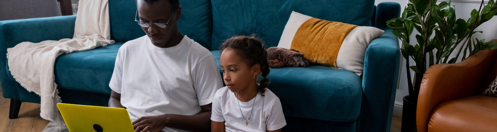 A man and a young girl sit together on the floor in a cosy living room, looking at a laptop together.