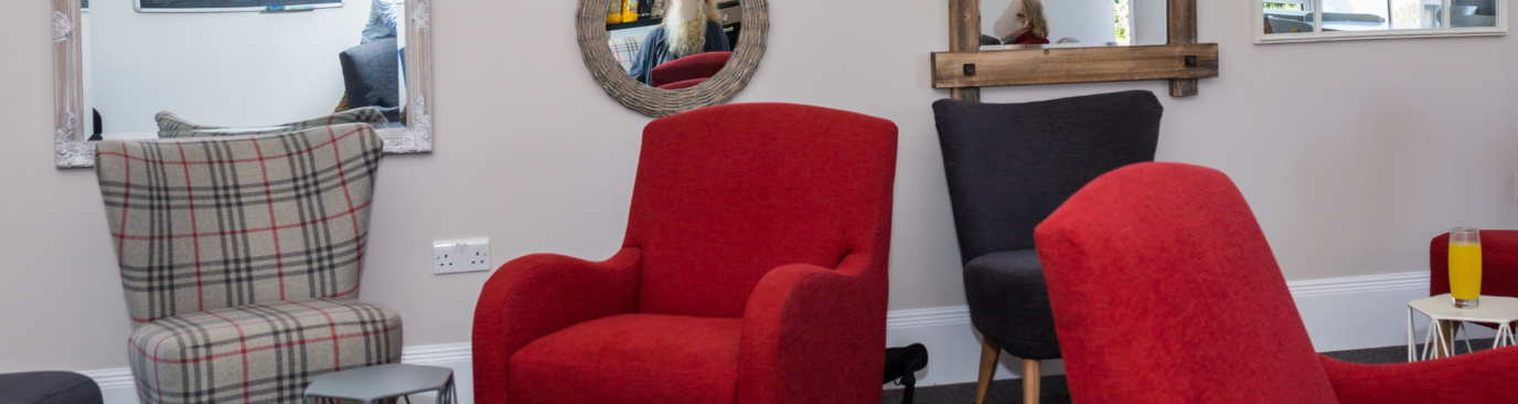 A welcoming lounge area with a mix of red and tartan armchairs arranged around decorative mirrors on neutral walls, with a glass of orange juice on a side table.