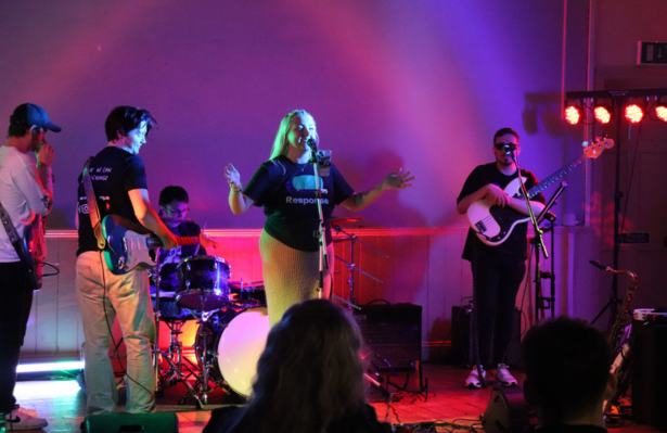A band performs on a colourfully lit stage at a fundraising gig, as a female speaks to an audience in the foreground.