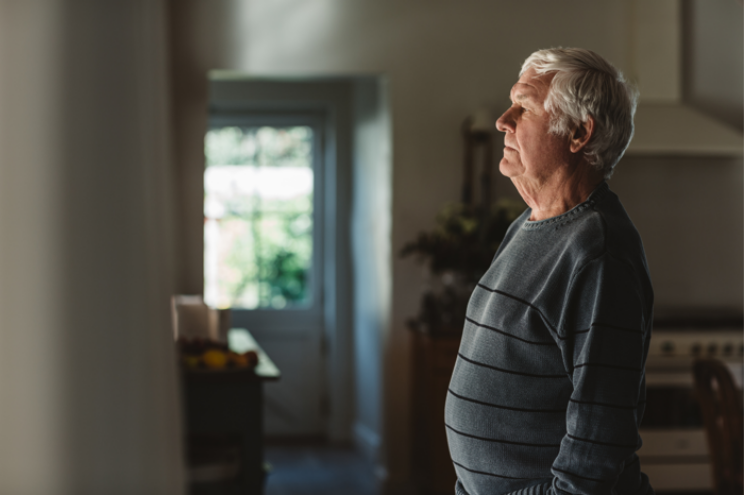 Elderly man in grey jumper stood inside, looking out of window.