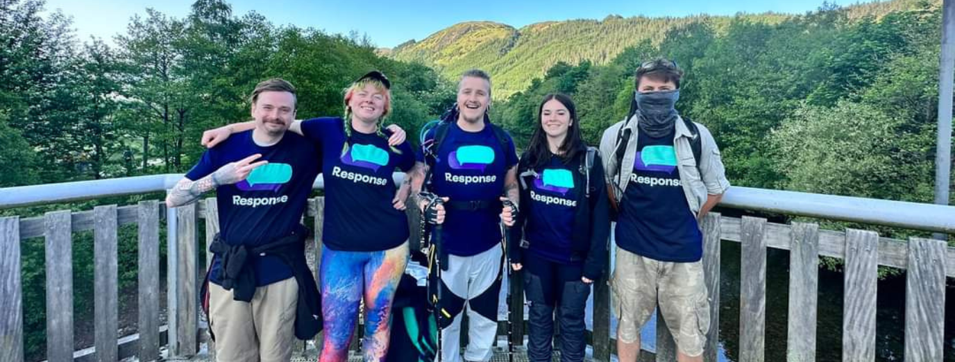 Five smiling fundraisers wearing Response t-shirts stand together on a bridge or viewpoint, with a forested hillside and blue sky behind them, equipped with walking gear for a charity hike.