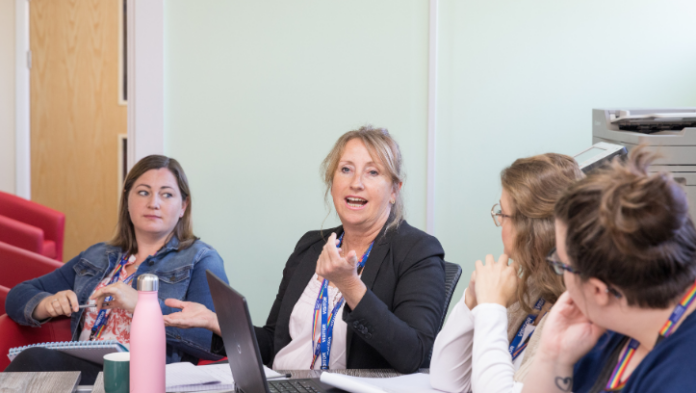A woman wearing a lanyard speaks attentively to colleagues during a professional meeting, with two other women listening around the table in an office setting.
