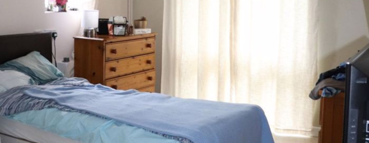 A comfortable single bedroom with a pine chest of drawers, blue bedding, and natural light coming through sheer curtains.