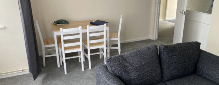 A simply furnished shared living space with a grey sofa and a light wood dining table with four white chairs.