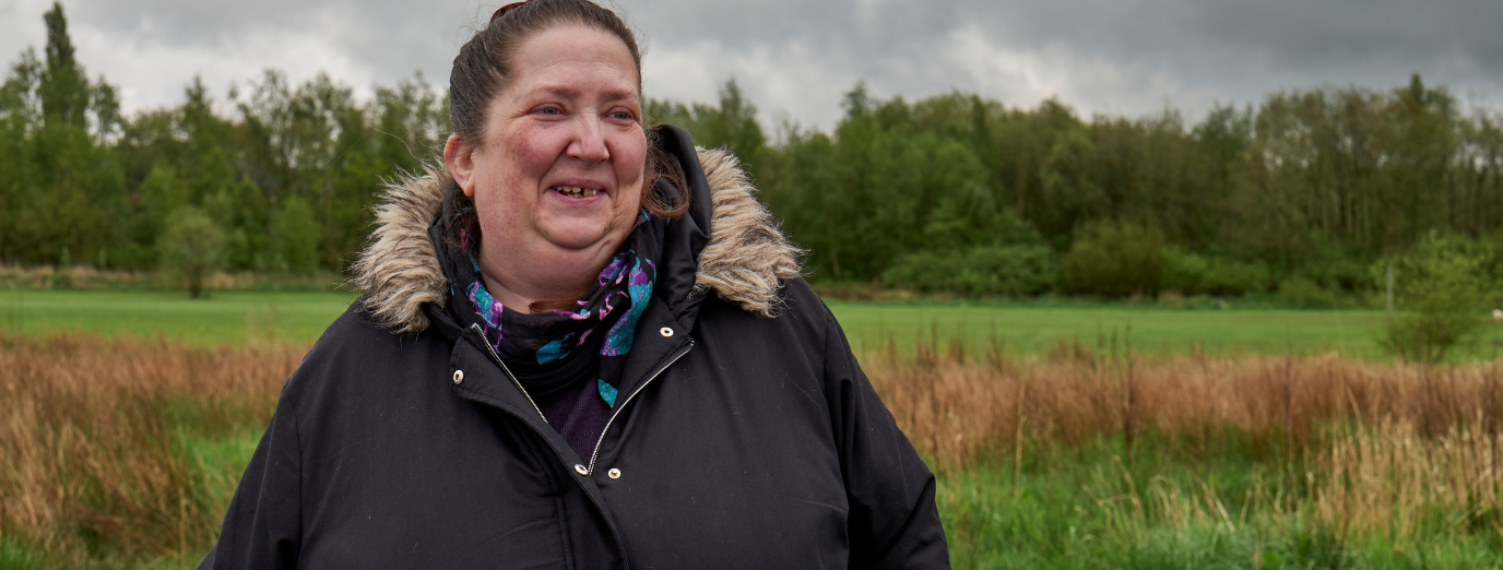 A woman in a dark winter coat and colourful scarf smiles warmly while standing outdoors in a green, open landscape on a cloudy day.