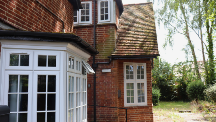 A red brick building with white-framed windows and a sloped tiled roof, surrounded by greenery and trees on a sunny day.