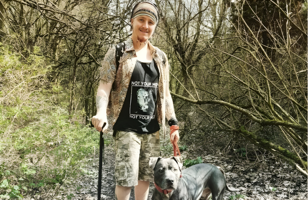 A person smiles while standing on a woodland trail holding a walking pole, with a grey dog on a lead beside them.