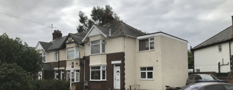 The exterior of a semi-detached residential house on a quiet street, with white rendered walls and a grey tiled roof on an overcast day.