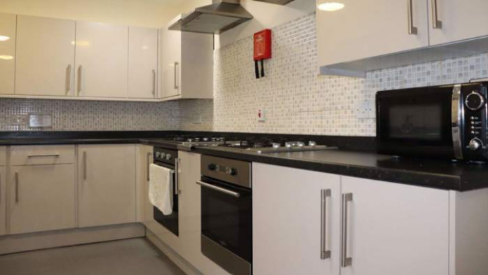 A clean, modern kitchen with white cabinets, two ovens, a stovetop, a microwave, and tiled backsplash, designed for shared use.